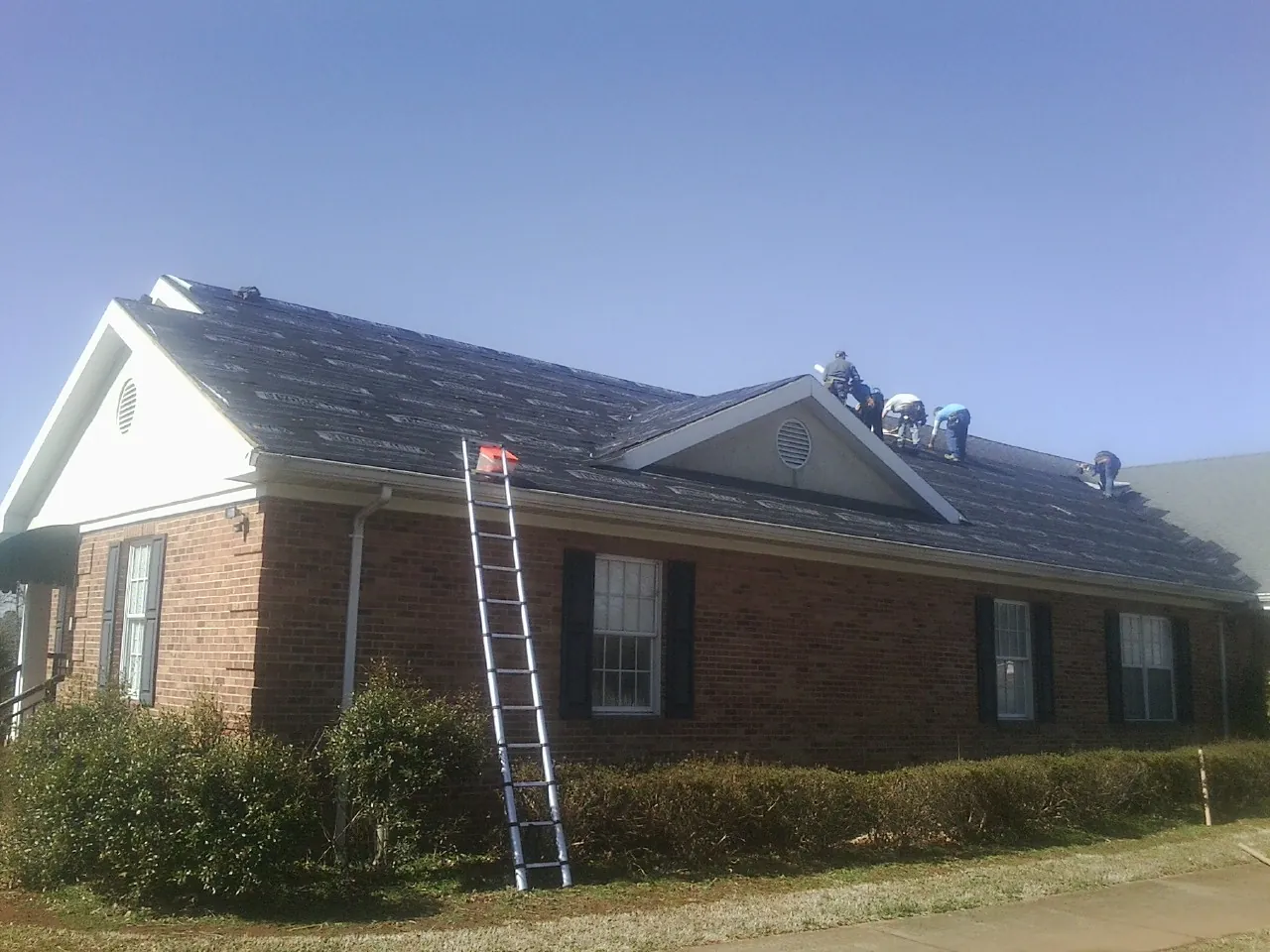 Crew tearing shingles off a brick ranch roof with a ladder set — Metal Roof Repair work by our crew in Kersey