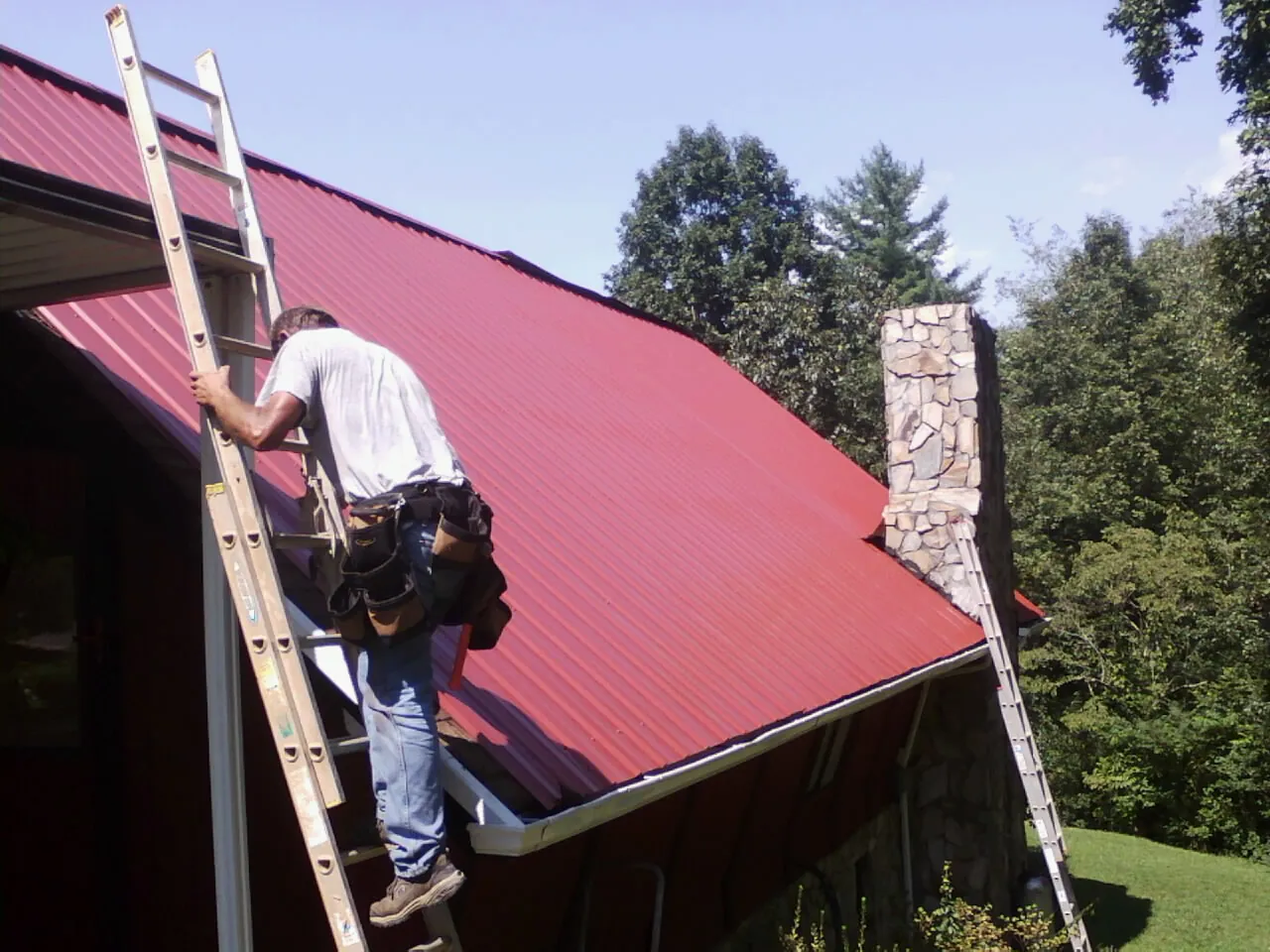 Roofer on a ladder beside a red metal roof and stone chimney — Soffit & Fascia Repair by Roofing Brothers in Kersey